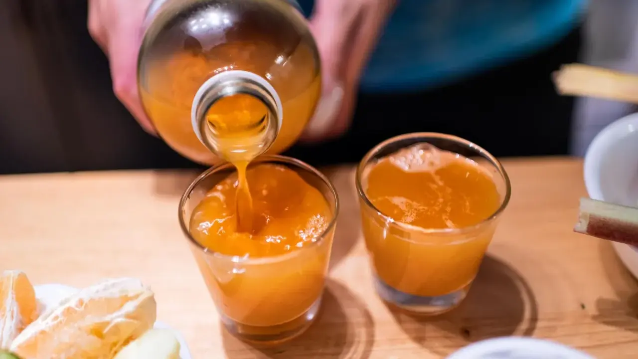 Przygotowanie domowego sposobu na detox wątroby. Pomarańczowy sok z butelki nalewany do szklanek, obok owoce.