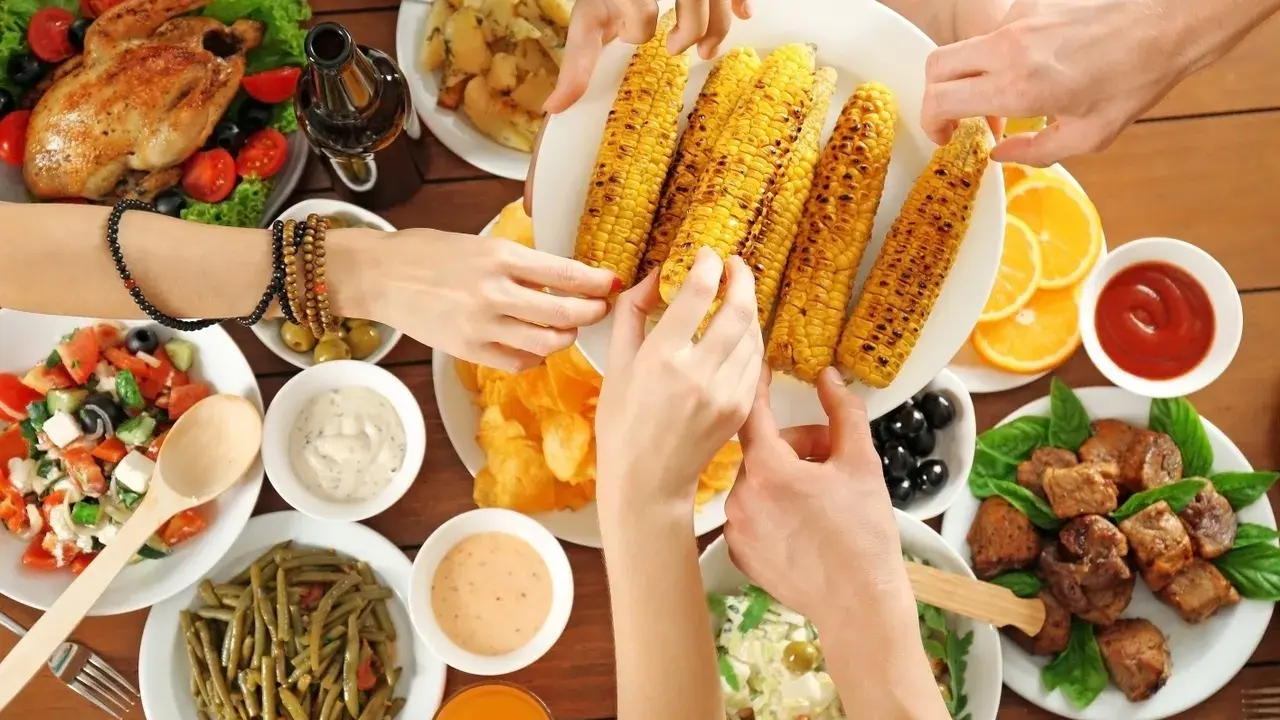 Ręce sięgają po grillowaną kukurydzę na keto. Na stole pieczony kurczak, sałatki, mięso i sosy.