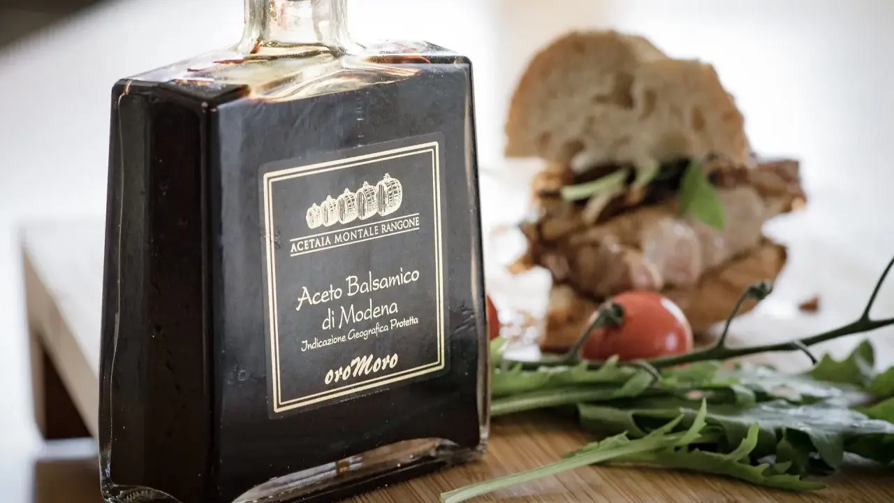 Butelka octu balsamicznego di Modena, z etykietą "Aceto Balsamico di Modena" i "oro Moro". W tle kanapka i rukola.