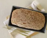 Świeżo upieczony chleb orkiszowy w keksówce, posypany ziarnami. Obok leżą kłosy zbóż.