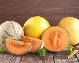 Kuszące plastry i połówka melona pomarańczowego w środku, obok innych melonów i listków mięty na drewnianym stole.