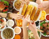 Ręce sięgają po grillowaną kukurydzę na keto. Na stole pieczony kurczak, sałatki, mięso i sosy.