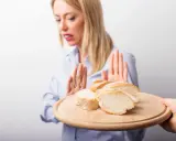 Kobieta z przerażeniem odrzuca talerz z kromkami chleba. Jej gesty sugerują uczulenie na chleb lub nietolerancję glutenu.