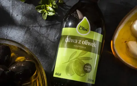 Butelka oliwy z oliwek Olini z Hiszpanii, obok miseczki z czarnymi oliwkami i ząbkami czosnku. Idealna do potraw, nawet tych wymagających gorzkiej oliwy z oliwek.
