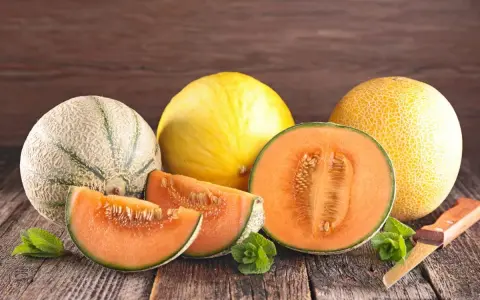Kuszące plastry i połówka melona pomarańczowego w środku, obok innych melonów i listków mięty na drewnianym stole.