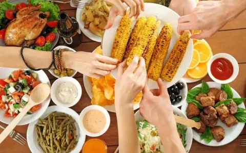 Ręce sięgają po grillowaną kukurydzę na keto. Na stole pieczony kurczak, sałatki, mięso i sosy.