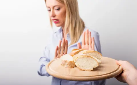 Kobieta z przerażeniem odrzuca talerz z kromkami chleba. Jej gesty sugerują uczulenie na chleb lub nietolerancję glutenu.
