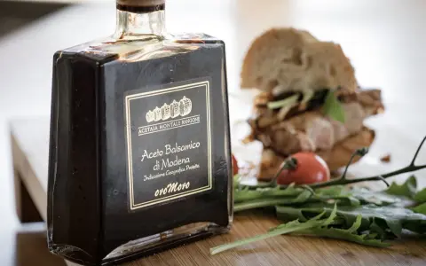 Butelka octu balsamicznego di Modena, z etykietą "Aceto Balsamico di Modena" i "oro Moro". W tle kanapka i rukola.