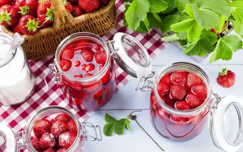 Słoiki z truskawkami w syropie, koszyk pełen owoców i cukiernica. Idealne na przetwory, gdy szukasz, czym zastąpić cukier żelujący.