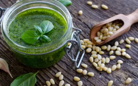 Słoik domowego pesto, świeża bazylia, czosnek i orzeszki piniowe na drewnianym blacie. Pesto co to? To aromatyczny sos idealny do makaronu.