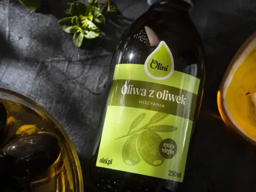 Butelka oliwy z oliwek Olini z Hiszpanii, obok miseczki z czarnymi oliwkami i ząbkami czosnku. Idealna do potraw, nawet tych wymagających gorzkiej oliwy z oliwek.