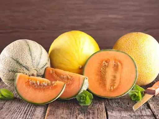 Kuszące plastry i połówka melona pomarańczowego w środku, obok innych melonów i listków mięty na drewnianym stole.