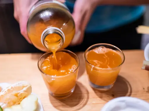Przygotowanie domowego sposobu na detox wątroby. Pomarańczowy sok z butelki nalewany do szklanek, obok owoce.