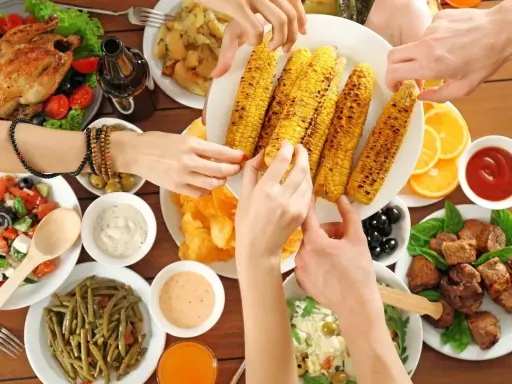 Ręce sięgają po grillowaną kukurydzę na keto. Na stole pieczony kurczak, sałatki, mięso i sosy.