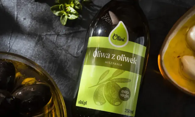 Butelka oliwy z oliwek Olini z Hiszpanii, obok miseczki z czarnymi oliwkami i ząbkami czosnku. Idealna do potraw, nawet tych wymagających gorzkiej oliwy z oliwek.