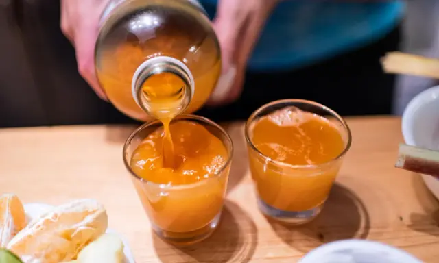 Przygotowanie domowego sposobu na detox wątroby. Pomarańczowy sok z butelki nalewany do szklanek, obok owoce.