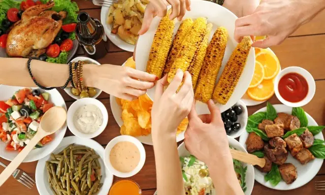 Ręce sięgają po grillowaną kukurydzę na keto. Na stole pieczony kurczak, sałatki, mięso i sosy.