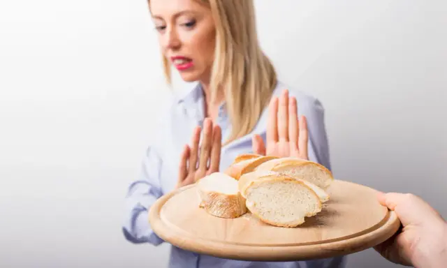 Kobieta z przerażeniem odrzuca talerz z kromkami chleba. Jej gesty sugerują uczulenie na chleb lub nietolerancję glutenu.